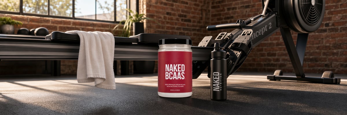 Naked BCAAs tub beside a rowing machine in a sunlit home gym with exposed brick walls.