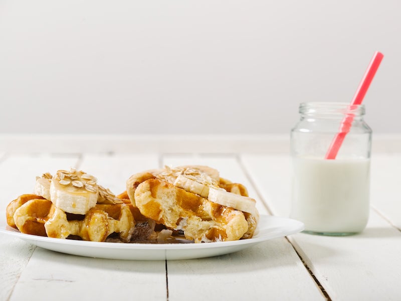 Waffles de proteína de plátano caseros en un plato, cubiertos con rodajas de plátano y almíbar con un vaso de leche al lado