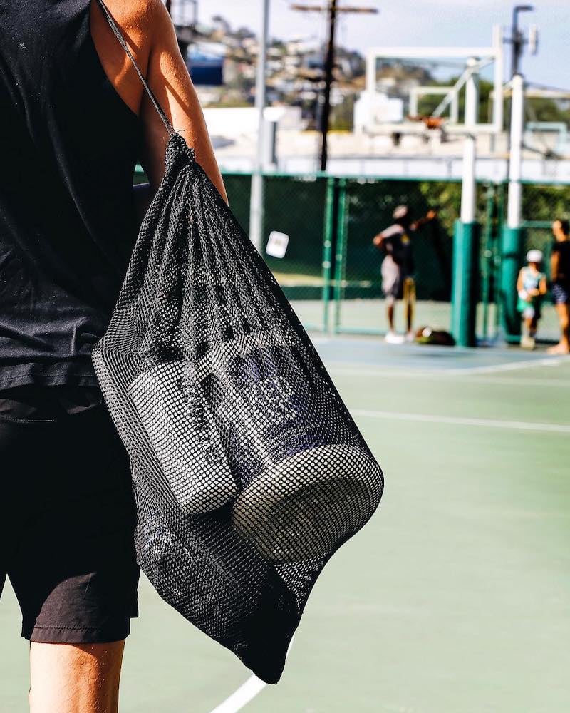 Un hombre camina por una cancha de tenis, llevando una bolsa de malla con un bote de Naked Mass y una botella mezcladora de Naked Nutrition