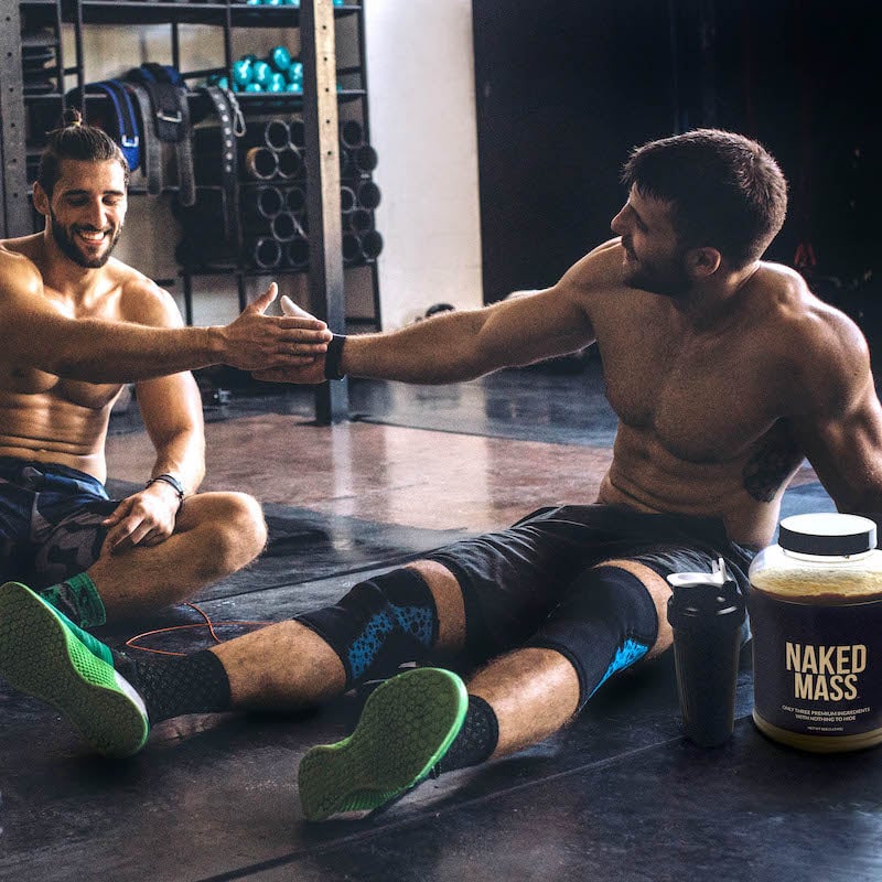Dos hombres sentados y descansando después de un entrenamiento en el gimnasio, una tina de Naked Mass está a su lado.