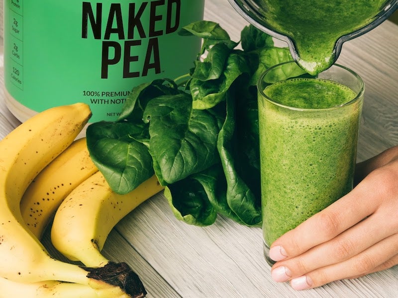 Imagen del producto Naked Pea con un bote del producto junto a un batido de proteína de guisante verde vertido en un vaso con hojas de espinaca y plátano.