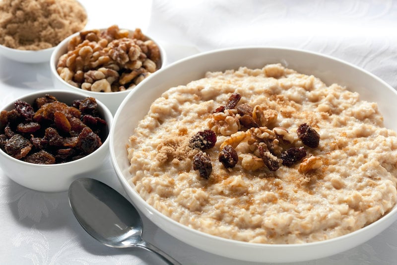 Avena cocida en un bol, junto a tazones más pequeños de pasas, nueces y azúcar morena.